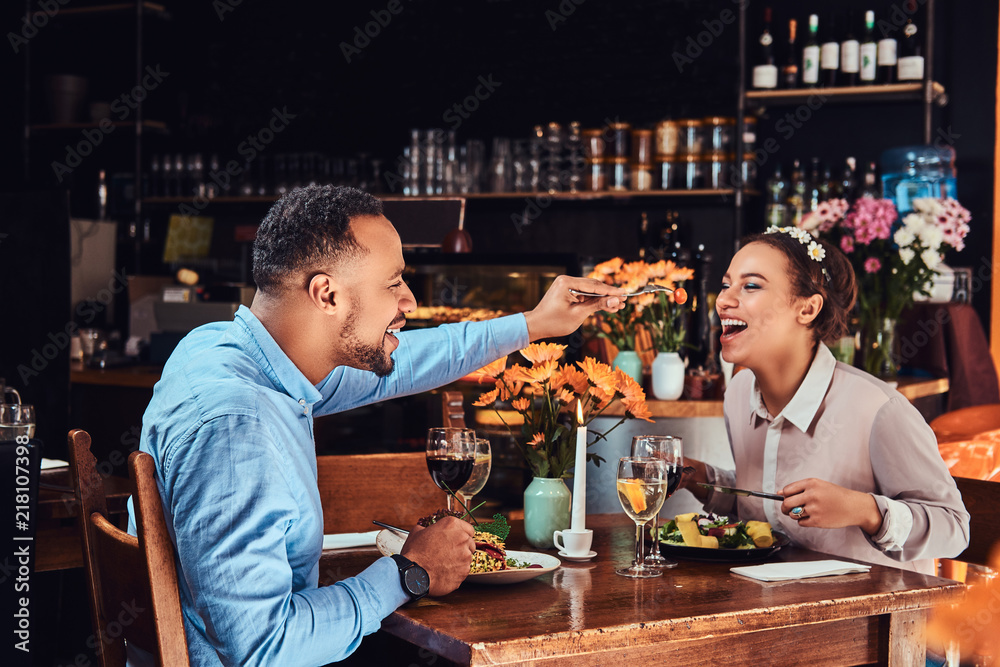 Beautiful African-American couple in love having a great time together at their dating, an attractive couple enjoying each other, handsome man feeding his woman in restaurant. Beautiful African-American couple in love having a great time together at their dating, an attractive couple enjoying each other, handsome man feeding his woman in restaurant.