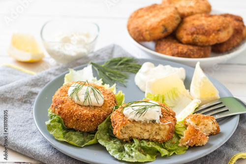 Salmon patties served on green lettuce leaves with fresh dill, lemon slices and white sauce on top.