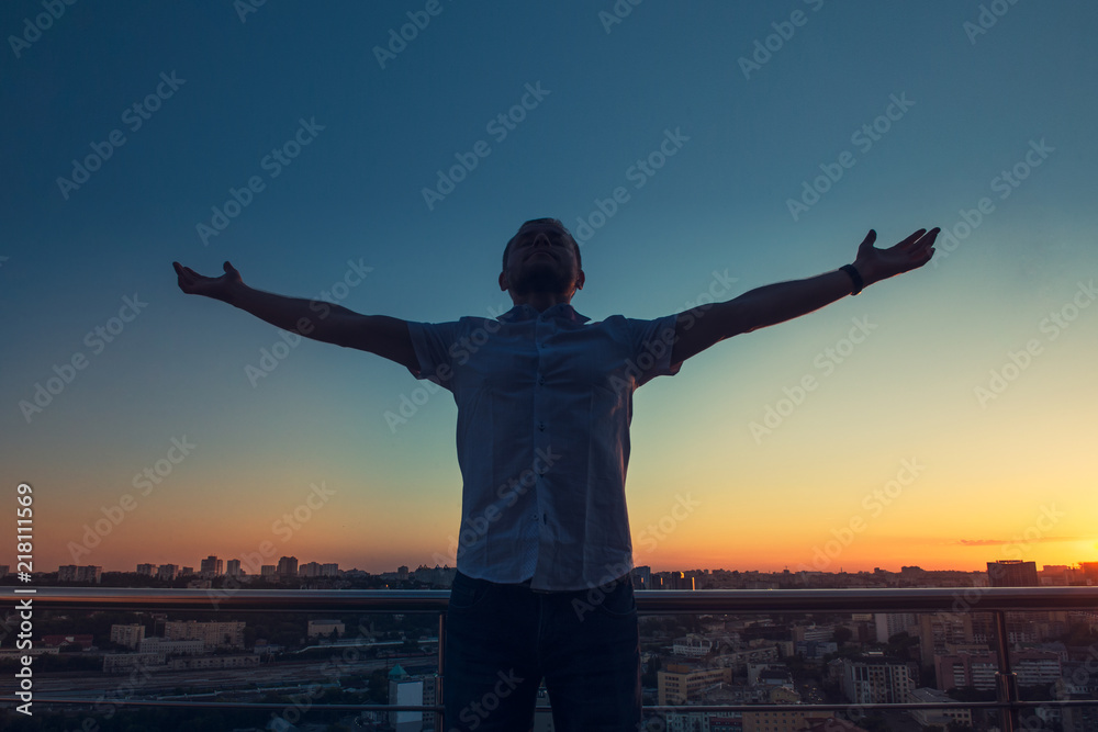 Man's silhouette with hands rised up to sky on sunset cityscape bacground. Feeling and celebrating freedom, victory, sucsess. Expressing joy of life. Positive emotion. Selective focus. Copy space. Man's silhouette with hands rised up to sky on sunset cityscape bacground. Feeling and celebrating freedom, victory, sucsess. Expressing joy of life. Positive emotion. Selective focus. Copy space.