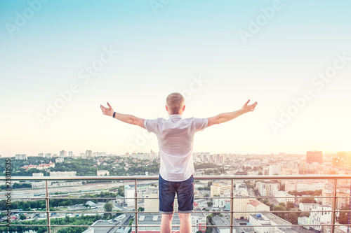 Back view Man on a high rise building balcony overlooking city with hands rised up to sky, feeling and celebrating freedom, victory, sucsess. Expressing his joy of life. Positive emotions. Copy space.