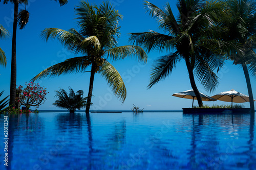 Vacation background with infinite swimming pool and palm trees. Blue ripped water in swimming pool with reflection green palm trees. Tropical resort pool with lounge chairs and ocean view.