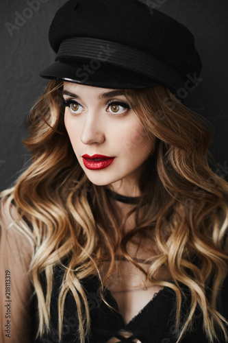 Glamorous and fashionable portrait of beautiful and sexy blonde model girl with bright makeup and with full sexy lips, in the black hat and in a stylish black lingerie posing at dark background