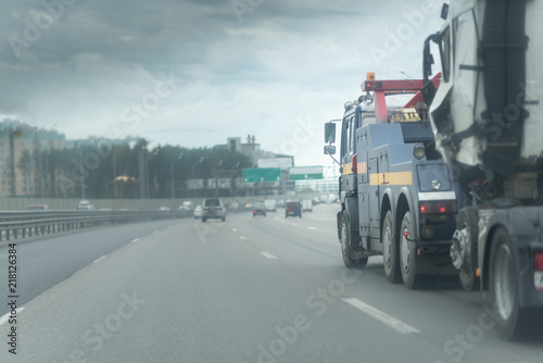 Blue Tow truck delivers the damaged white lorry, Sun light flare, Selective focus. Asphalt highway with metal safety barrier or rail. Clouds at the sky