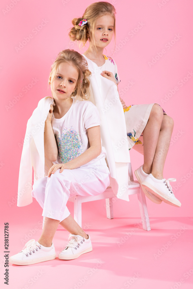 Twins girls in bright clothes posing near the stairs with two steps on a pink background. Twins girls in bright clothes posing near the stairs with two steps on a pink background.