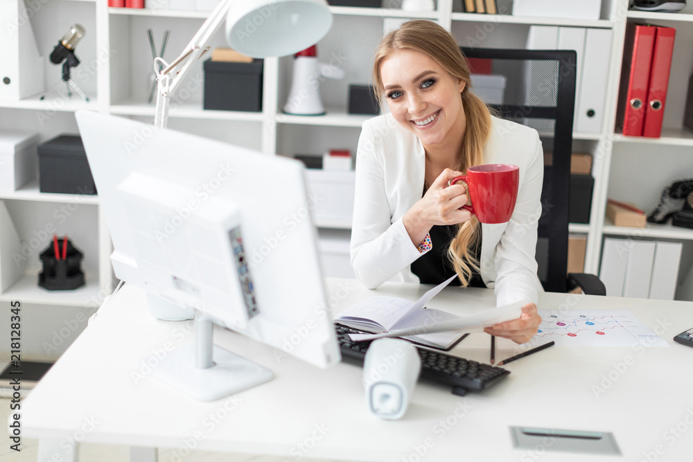 A young girl is sitting at the computer desk in the office and is holding a cup and documents. Before the girl on the table are diagrams. A young girl is sitting at the computer desk in the office and is holding a cup and documents. Before the girl on the table are diagrams.
