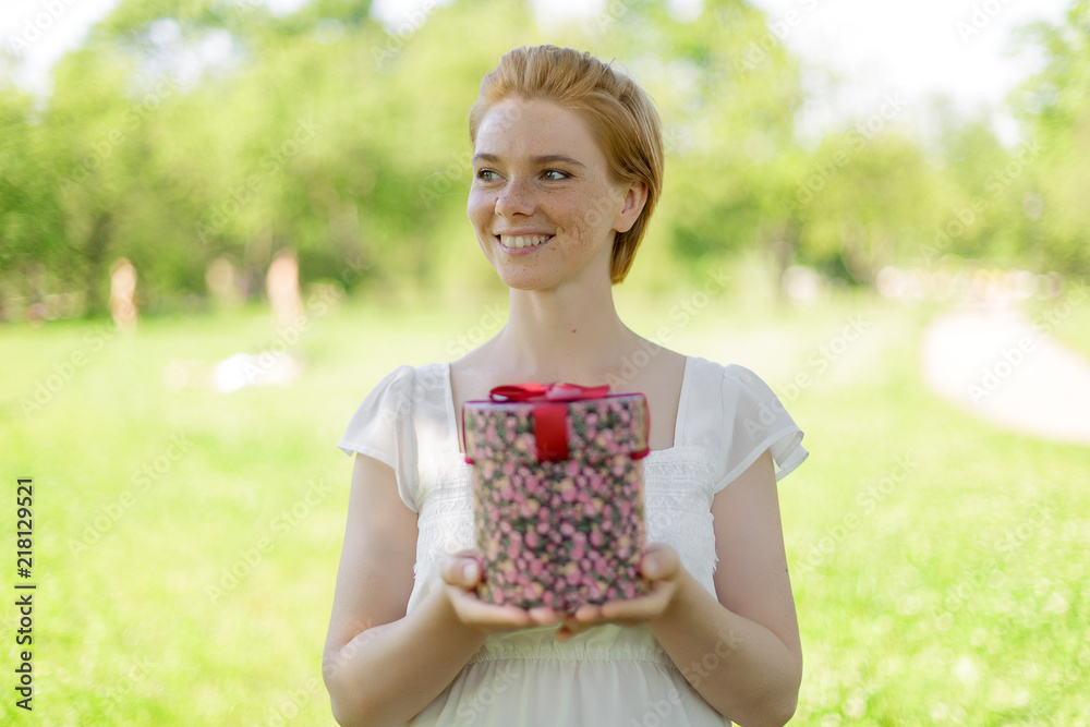 This is for you. Attractive young woman holding a gift box in her hands standing against summer green park