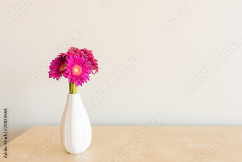 Fototapeta Naklejka Na Ścianę i Meble -  Closeup of bright pink gerbera daisies in small white vase on wooden shelf against neutral wall background (selective focus)