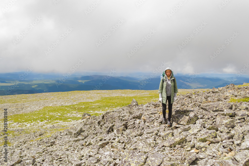 A girl in a green jacket with a hood with a backpack stands on the rocks rising in the mountains of the Altai against the background of dense clouds lying on the peaks of the hills