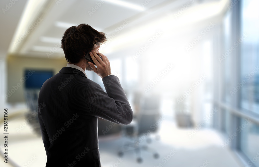 Obraz premium Businessman standing in the office talking with his cell phone. Sunlight enters by for the window.