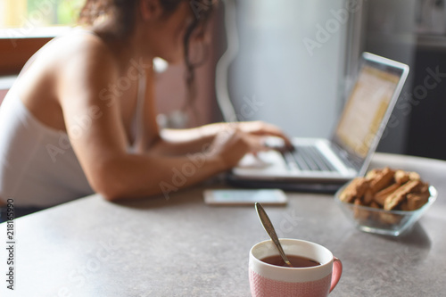Cup of tea on grunge kitchen table and woman sitting and using laptop on blurred background. Freelancer works at home online marketing, education e-learning concept. Sdf with focus at cup with focus a