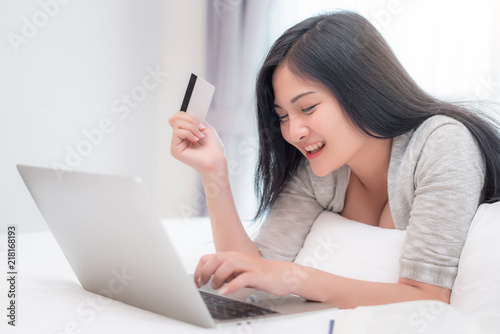 Pretty attractive Asian woman shopping online by laptop with credit card, laying on the bed at bedroom 