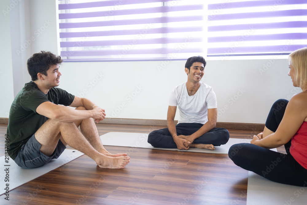 Students talking with smiling instructor after yoga class. Men and woman sitting on mats in gym. Yoga class concept.