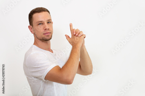 Confident young male super-agent with stubble making finger gun and looking at camera. Serious man in t-shirt gesturing against white background. Brave man concept photo