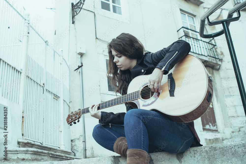 Ταπετσαρία Closeup portrait of focused young beautiful woman playing guitar and sitting on city stairs with building in background