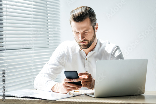 handsome fashionable businessman using smartphone at workplace with laptop and documents