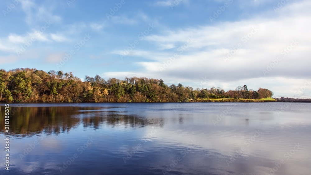 A tree covered peninsula on a bright sunny day with reflections on a calm lake.