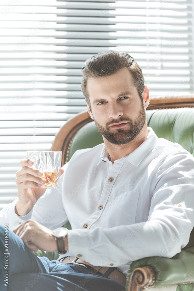 confident stylish man holding glass of whiskey and sitting in armchair