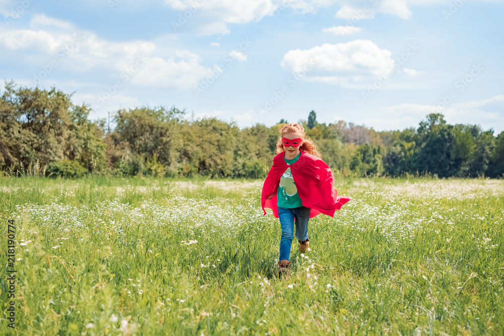 little kid in red superhero cape and mask running in meadow on summer day little kid in red superhero cape and mask running in meadow on summer day