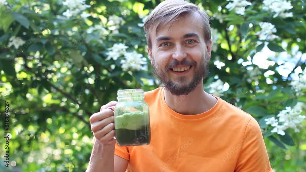 Handsome man drinks fresh green smoothie from jar and smile in summer garden. Healthy food or vegan concept. Handsome man drinks fresh green smoothie from jar and smile in summer garden. Healthy food or vegan concept.