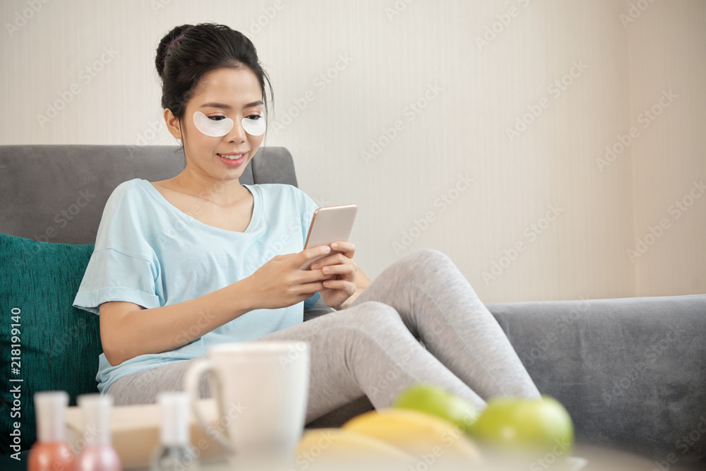 Charming Asian female with under-eye patches smiling and using smartphone while sitting on comfortable sofa in living room Charming Asian female with under-eye patches smiling and using smartphone while sitting on comfortable sofa in living room
