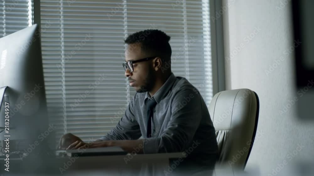 Side view of tired black man in glasses writing texts using desktop computer when sitting at desk in office Side view of tired black man in glasses writing texts using desktop computer when sitting at desk in office
