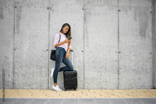 Woman using smart phone while leaning on the luggage on the street.