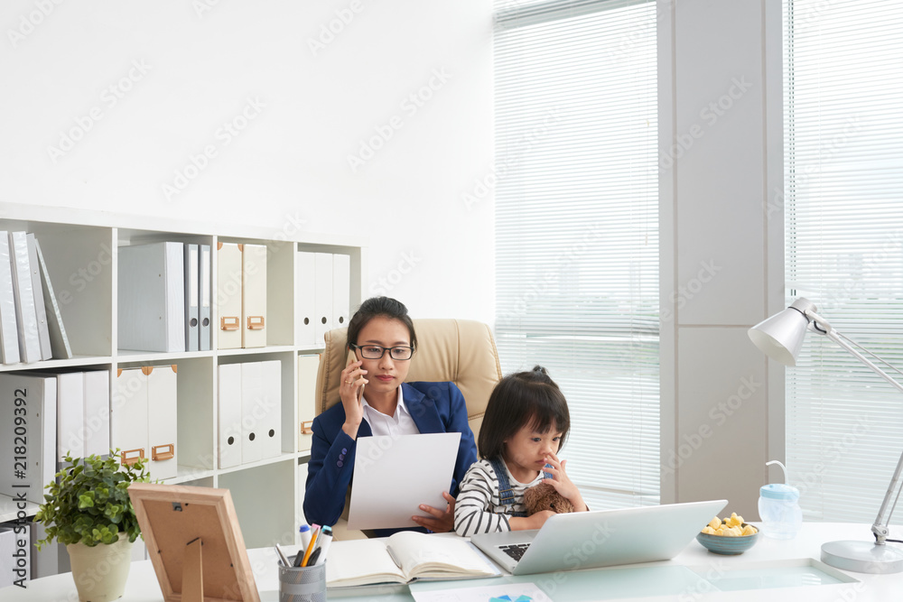Adult businesswoman having phone call and looking papers with adorable daughter sitting on knees at workplace