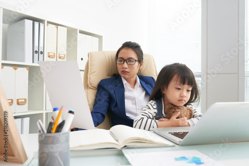 Asian woman in suit reading document while working in office with charming little daughter