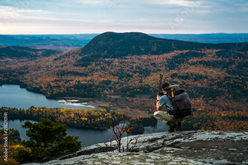 Leinwand Poster A man kneels on a mountain in Maine overlooking a lake and fall foliage while hi