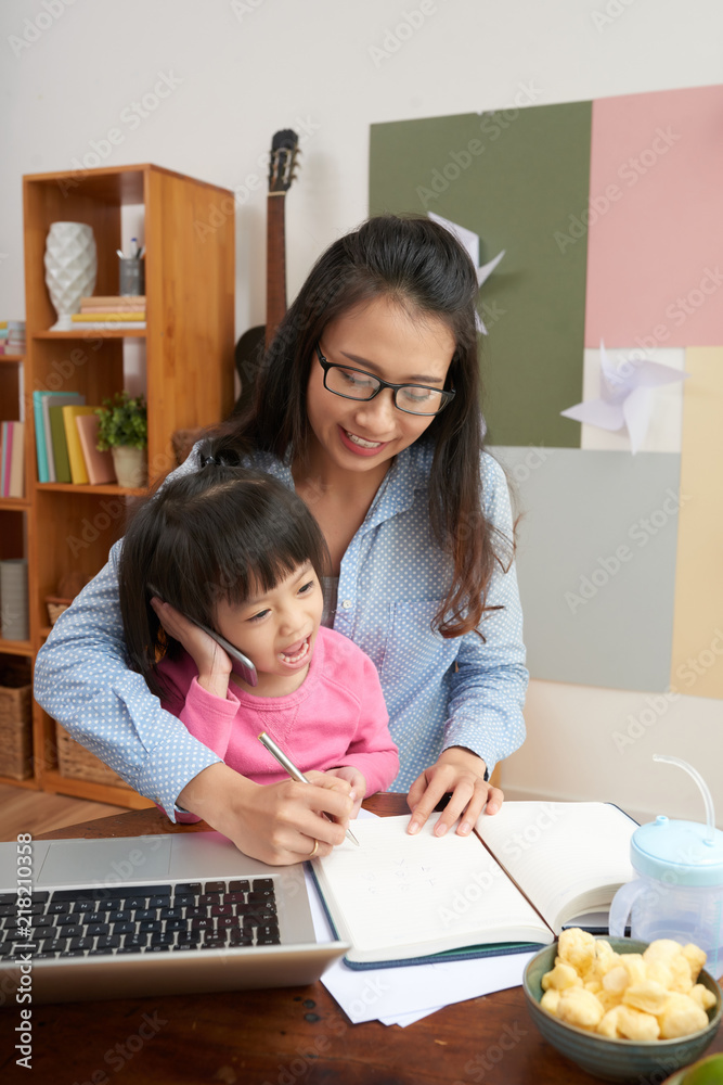 Pretending little Asian girl with smartphone sitting at table with adult mother working with laptop and writing in notepad Pretending little Asian girl with smartphone sitting at table with adult mother working with laptop and writing in notepad