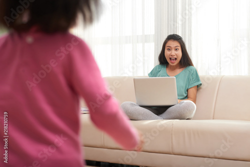 Asian emotional woman sitting with laptop on sofa and looking excited with view of running little daughter 