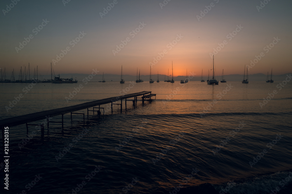Beautiful view of wooden pier and sailing boats at the calm sea on pink and orange sunset. Mountains on background. Twilight