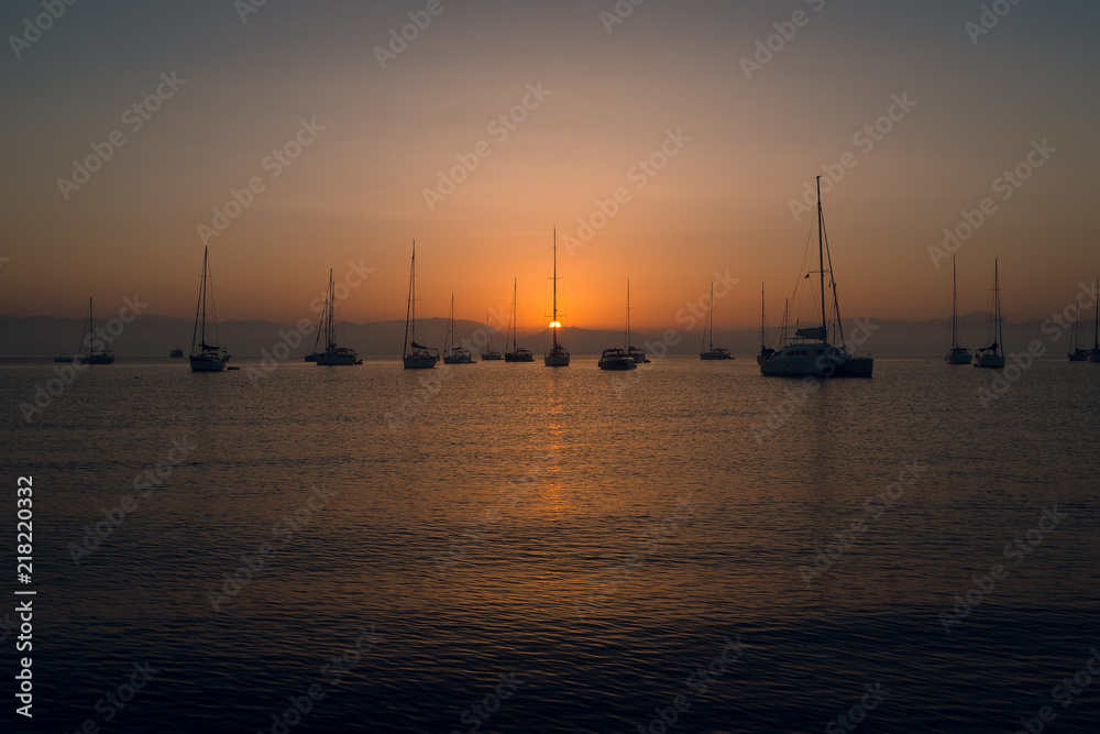 Yachts and boats at the calm sea bay with mountains on background at beautiful orange sunset. Dusk. Greece