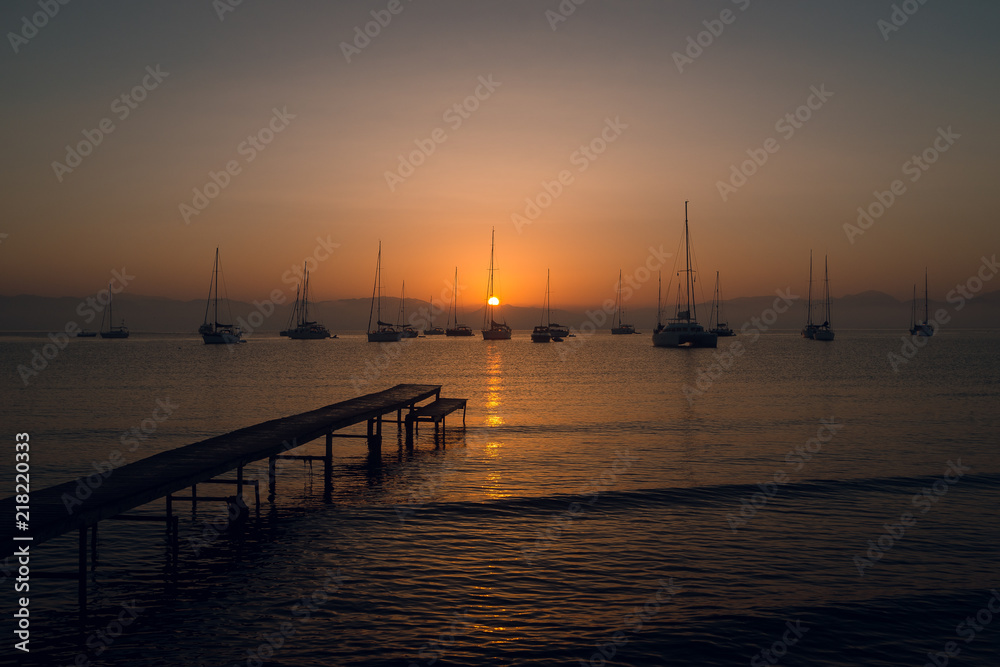 Wooden pier, yachts and boats at the calm sea bay with mountains on background at beautiful orange sunset. Dusk. Greece