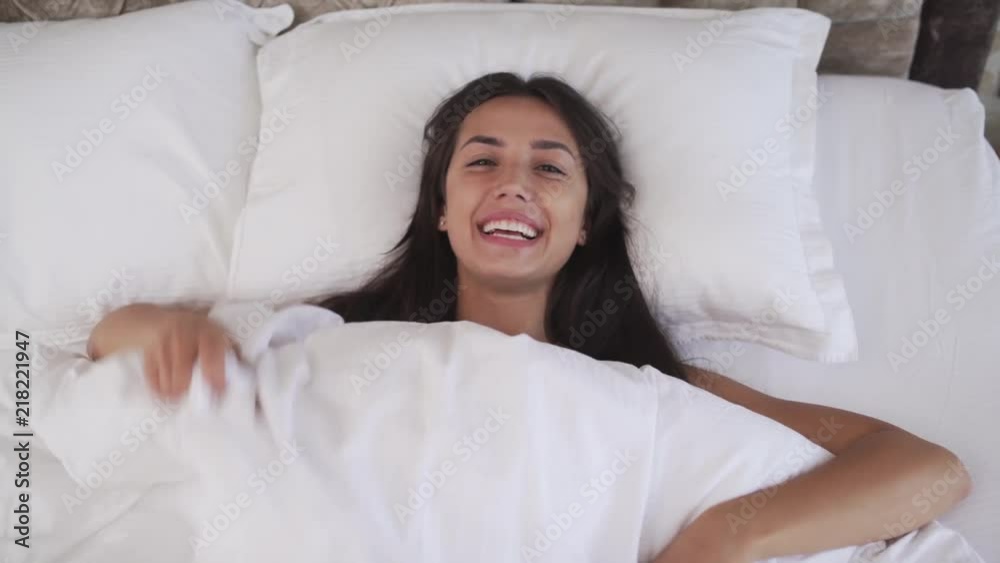 A top view of a young brunette hiding under the blanket. She throws away the blanket, smiles at the camera, sticks the tongue, makes happy faces, peeps and winks lying on the bed. A top view of a young brunette hiding under the blanket. She throws away the blanket, smiles at the camera, sticks the tongue, makes happy faces, peeps and winks lying on the bed.