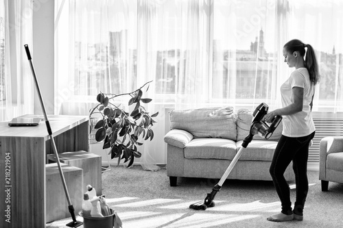 Full body portrait of young woman in white shirt and jeans cleaning carpet with vacuum cleaner in living room, copy space. Housework, cleanig and chores concept photo