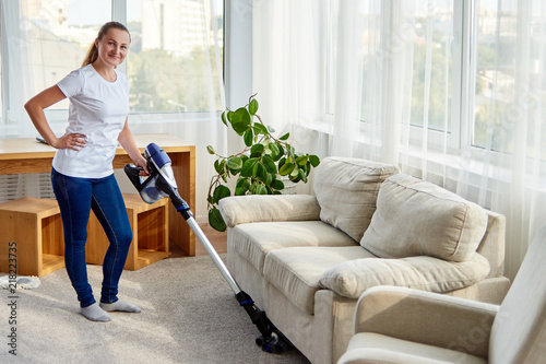 Full body portrait of young woman in white shirt and jeans cleaning carpet with vacuum cleaner in living room, copy space. Housework, cleanig and chores concept