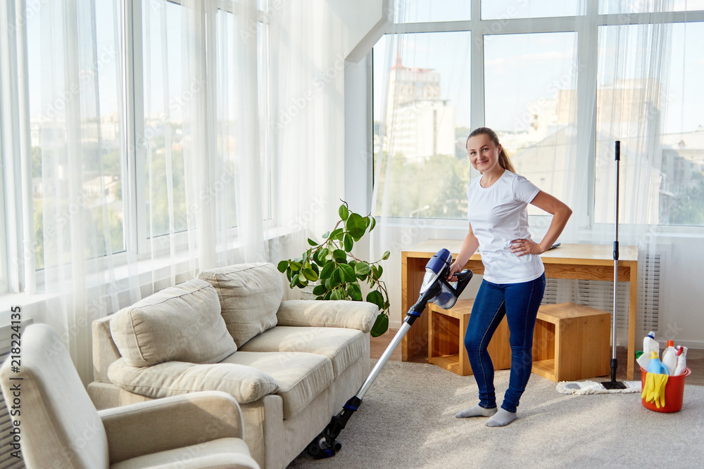 Full body portrait of smiling woman in white shirt and jeans cleaning carpet with vacuum cleaner in living room, copy space. Housework, cleanig and chores concept Full body portrait of smiling woman in white shirt and jeans cleaning carpet with vacuum cleaner in living room, copy space. Housework, cleanig and chores concept