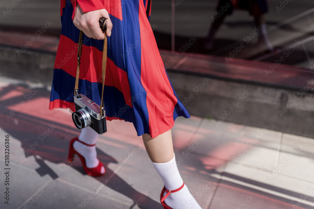 Obraz premium Elegant lady wearing stylish red skirt and shoes holds in hands vintage photo camera. Low angle view of her shapely legs. Selective focus