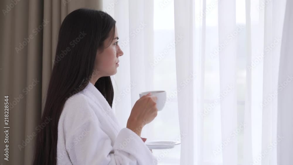 A close up of a young brunette drinking tea or coffee by the window. Light wind moves the curtains. She makes one sip, tastes it, bends head, sighs, and keeps looking through the window. A close up of a young brunette drinking tea or coffee by the window. Light wind moves the curtains. She makes one sip, tastes it, bends head, sighs, and keeps looking through the window.
