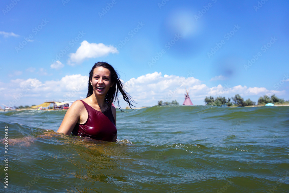 Young brunette girl in red swimsuit smiling and swimming in a sea, with travel camp and indian teepees wigwams on a beach background and blue cloudy sky. Copyspace.