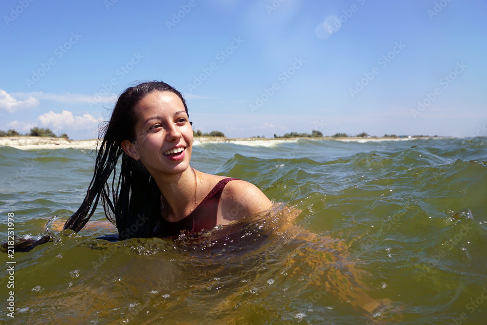 Young brunette girl in red swimsuit smiling and swimming in a sea, with sandy beach island background and  blue sky. Copyspace.
