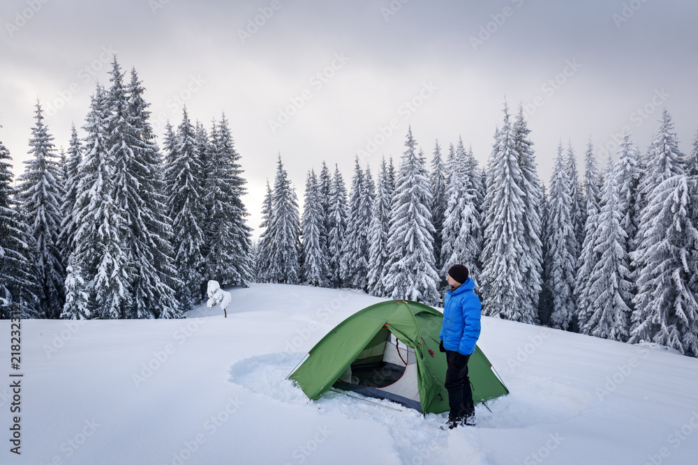 Green tent and tourist against the backdrop of snowy pine tree forest. Amazing winter landscape. Tourists camp in high mountains. Travel concept