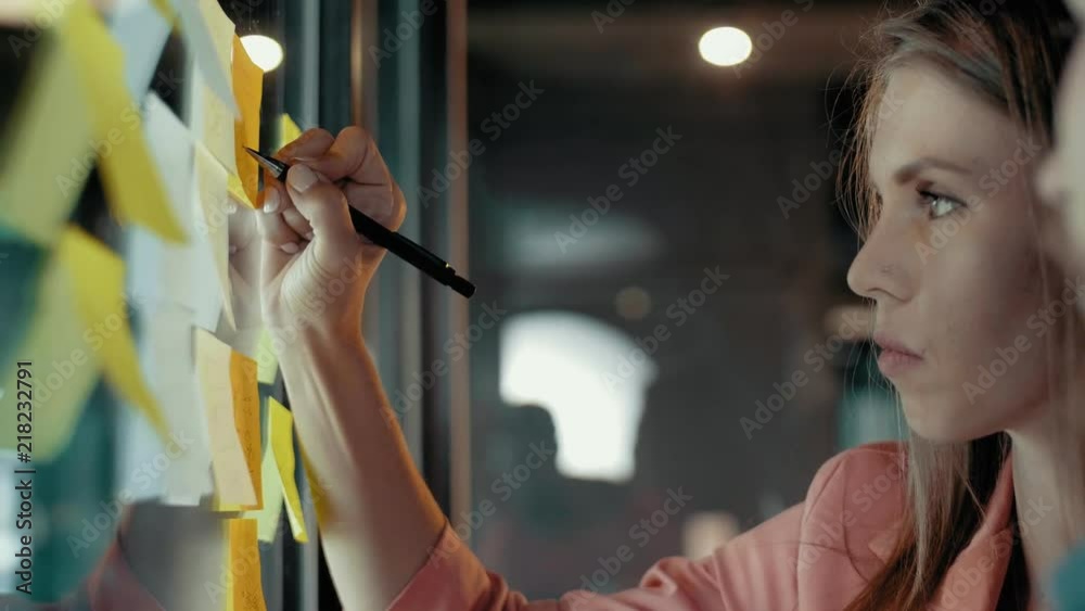 Side view multiethnic creative business team meeting brainstorming analyzes concept and planning strategy of start up project look at transparent wall with sticky multicolored stickers and take notes