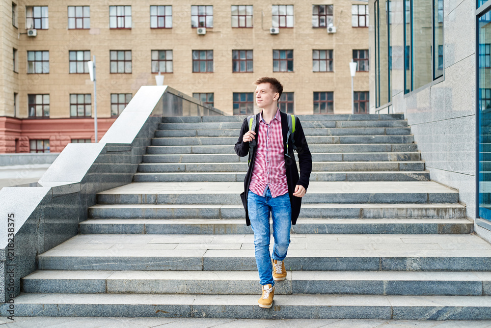 Full length body portrait of handsome male student with backpack walking down stairs and having rest in university campus  outdoors, copy space. Education, rest and relax, travel and tourism concept