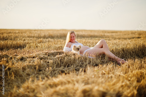 Blonde pregnant woman in wreath field at white underwear clothes with dog on sunset. Happy moments of pregnancy.
