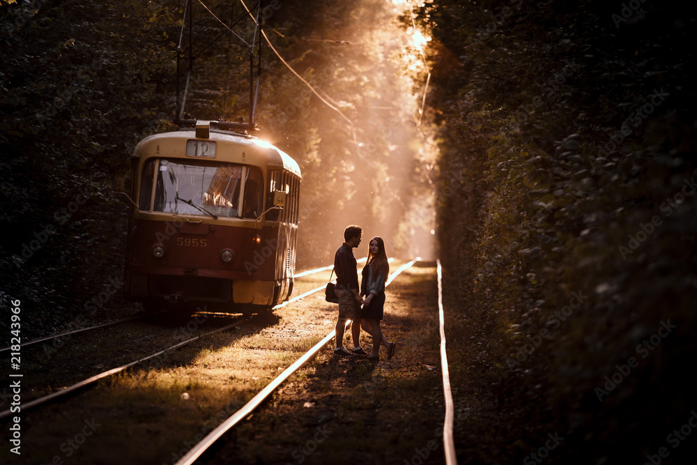 Naklejka premium Young couple, standing on the tram rails, in high trees alley in stunning evening sunlight.