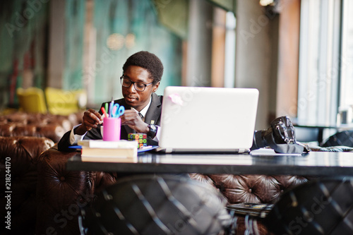Business african american man wear on black suit and glasses sitting at office with laptop and working, pours sugar into coffee.