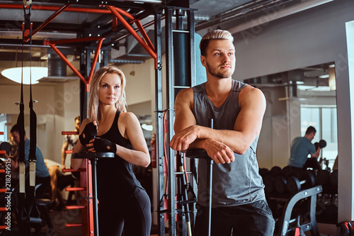 Portrait of an attractive sportive couple posing while leaning on barbells, looking away in a fitness club or gym.