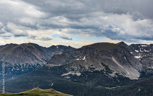 Clear, blue-gray Colorado Rocky Mountains, with dark evergreen forest in front, and two-pointed green rise in left-front.  Hint of storm clouds overhead.  Dramatic.  Good for background, text. © miles before I sleep